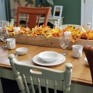 May include: A wooden box centerpiece with fall leaves and white candles sits on a table set with white plates, bowls, and mugs. The table is surrounded by white chairs.