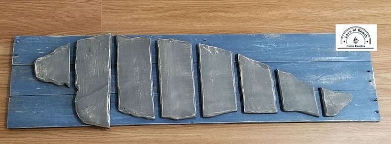 Puede incluir: Un letrero de madera con fondo azul y una forma de pez gris hecha de piezas individuales de madera. El letrero tiene el texto "Love of Beach Home Designs" en un c&iacute;rculo.