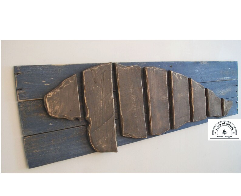 Puede incluir: Una pieza de arte de pared de madera con forma de concha, hecha de tablones de madera recuperados en tonos marrones y azules. La pieza est&aacute; montada sobre un fondo de madera pintado de azul. El texto "Love of Beach Home Designs" es visible en la esquina inferior derecha.