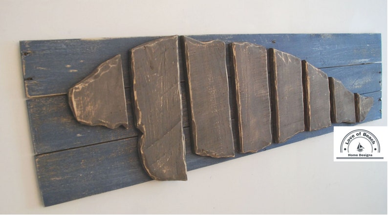 Puede incluir: Una pieza de arte de pared de madera con forma de concha, hecha de tablones de madera recuperados. La concha est&aacute; pintada de marr&oacute;n y montada sobre un fondo de madera pintado de azul. La pieza est&aacute; firmada con "Love of Beach Home Designs".