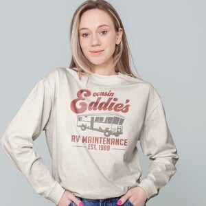 May include: A white long-sleeved sweatshirt with a vintage-style graphic design. The design features a red and white illustration of a camper van with the text "cousin Eddie's RV Maintenance Est. 1989".