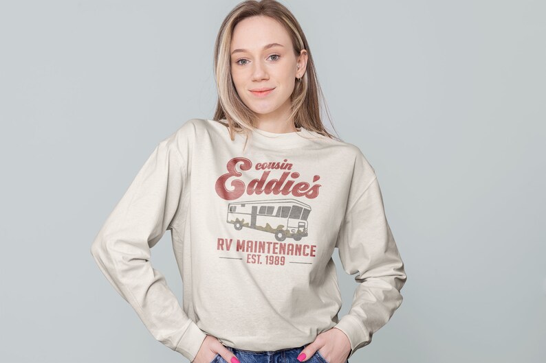 May include: A white long-sleeved sweatshirt with a vintage-style graphic design. The design features a red and white illustration of a camper van with the text "cousin Eddie's RV Maintenance Est. 1989".