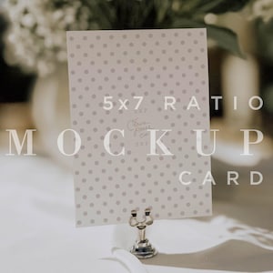 Op de afbeelding: Een mockup kaart met een 5x7 verhouding, met een lichtgrijs stippenpatroon. De kaart wordt vastgehouden door een zilverkleurige metalen standaard. De tekst "MOCK UP CARD" is zichtbaar. Witte bloemen op de achtergrond.