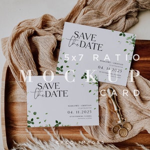 May include: A mockup of a save the date card with a 5x7 ratio. The card is white with a light blue background and features a green leafy design. The text on the card reads "Save the Date" and includes the names "Marlowe & Cristian" and the date "04.11.2025". The card is sitting on a wooden tray with a piece of beige fabric and a gold wax seal.