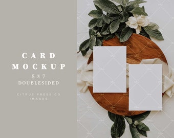 May include: Two blank 5x7 inch cards on a wooden tray with green leaves and white flowers. The cards are double-sided and perfect for mockups.