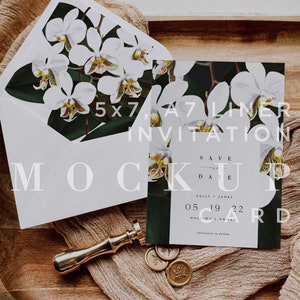 Op de afbeelding: Een witte envelop met een bloemenontwerp aan de voorkant, een witte A7-liner uitnodiging met een bloemenontwerp, een gouden waszegel en een houten dienblad met een bruine stof erop. De uitnodiging zegt "Save the Date" met de datum "05 19 22" en de locatie "Halawa, Hawaii".