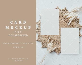May include: Two blank white 5x7 inch cards on a woven straw placemat with dried white pampas grass on a white background.  Card mockup.