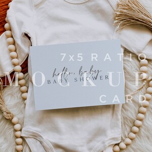 Pu&ograve; includere: Un body bianco per neonato con una carta celeste sopra. La carta ha il testo "7x5 Ratio Hello, Baby Baby Shower Card". Il body e la carta sono su una superficie bianca e soffice con una ghirlanda di perline di legno.