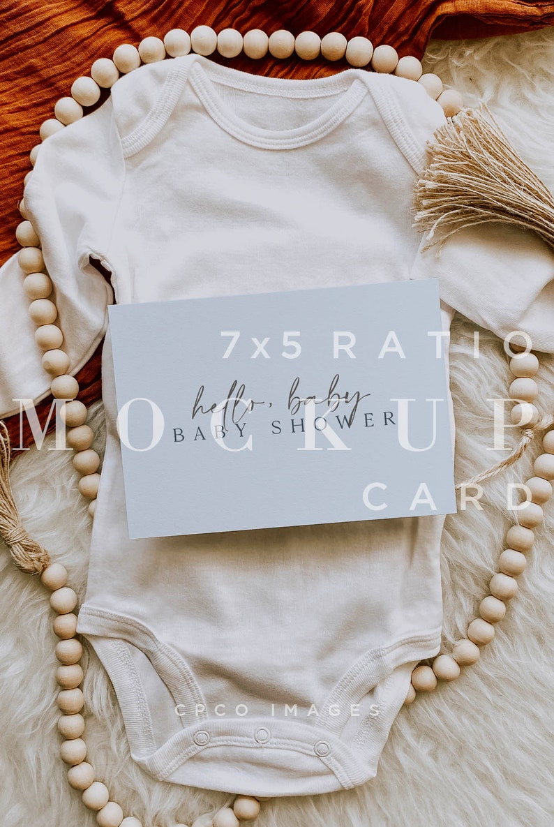 Pu&ograve; includere: Un body bianco per neonato con una carta celeste sopra. La carta ha il testo "7x5 Ratio Hello, Baby Baby Shower Card". Il body e la carta sono su una superficie bianca e soffice con una ghirlanda di perline di legno.