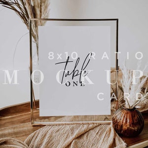 May include: A gold frame with an 8x10 ratio table number card mockup. The card is white with black text that says "table one". The frame is sitting on a wooden tray with a brown fabric runner and a small vase with dried pampas grass.