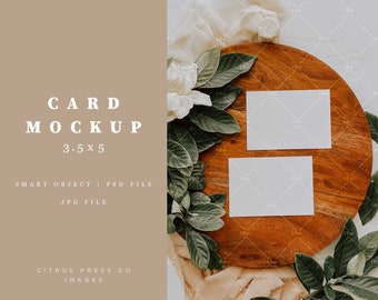May include: Two blank white cards on a wooden surface with green leaves and white flowers. The cards are 3.5 x 5 inches.