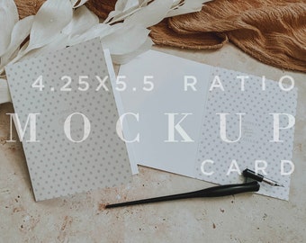 May include: A white card with a gray polka dot pattern and the text "4.25 x 5.5 Ratio Mockup Card" on it. The card is lying on a beige surface with a black calligraphy pen and a brown fabric with white dried grass.