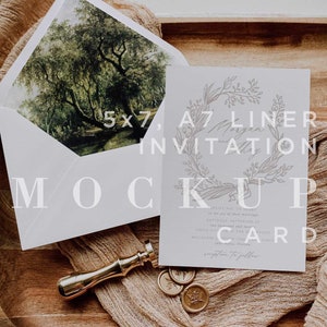 Op de afbeelding: Een witte uitnodigingskaart met een ontwerp van een bloemkrans en de tekst "A7 Liner Invitation". De kaart ligt op een houten dienblad met een gouden waszegel en een stuk beige stof.