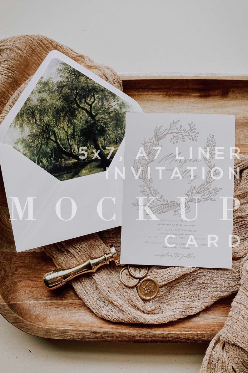 Op de afbeelding: Een witte uitnodigingskaart met een ontwerp van een bloemkrans en de tekst "A7 Liner Invitation". De kaart ligt op een houten dienblad met een gouden waszegel en een stuk beige stof.