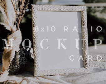 May include: A white picture frame with a textured border sits on a white fabric surface. The frame is empty and has an 8x10 ratio. The text "MOCKUP CARD" is printed below the frame. A bouquet of dried pampas grass and a gold ribbon are in the foreground.