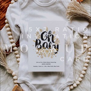 Pu&ograve; includere: Un body bianco con un invito per una baby shower di 12,7 x 17,8 cm sopra. La carta d'invito presenta un motivo floreale dorato e il testo "Oh Baby" in nero. Il testo "Unisciti a noi per una baby shower in onore di Ariel Carter" &egrave; stampato sotto il motivo floreale. La data e l'ora della festa sono stampate sotto il nome. Il body e la carta d'invito sono su una superficie marrone testurizzata.