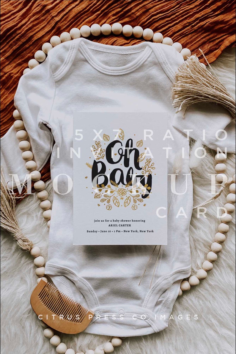 Pu&ograve; includere: Un body bianco con un invito per una baby shower di 12,7 x 17,8 cm sopra. La carta d'invito presenta un motivo floreale dorato e il testo "Oh Baby" in nero. Il testo "Unisciti a noi per una baby shower in onore di Ariel Carter" &egrave; stampato sotto il motivo floreale. La data e l'ora della festa sono stampate sotto il nome. Il body e la carta d'invito sono su una superficie marrone testurizzata.