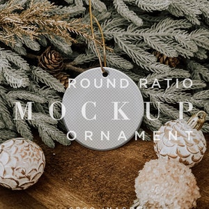 May include: A white round ceramic ornament with the text "ROUND RATIO MOCKUP ORNAMENT" on it. The ornament is hanging from a gold string and is surrounded by pine branches and white and gold ornaments.