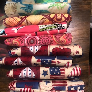 May include: A stack of fabric-covered items with various patterns. Designs include seahorses, turtles, seashells, geometric shapes, and patriotic themes with hearts, stars, and American flags. The color palette includes blues, greens, pinks, reds, and creams.