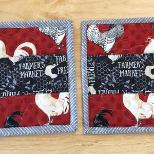 May include: Two square quilted pot holders with a red background featuring white rooster illustrations. A black and white patterned band with "FARMER'S MARKET FRESH" text is centered. The edges are trimmed with a gray and white striped fabric.