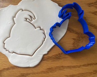 May include: A blue plastic cookie cutter in the shape of a rooster is next to a piece of white dough with the rooster shape pressed into it. The cookie cutter is on a wooden surface.