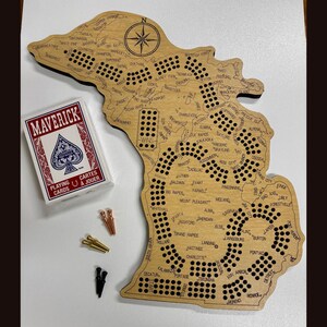 May include: A wooden map of Michigan with a game board design. The map is brown and has black text and black dots. The text includes city names and the word "Wins". The map is surrounded by a black border. A deck of playing cards is in the lower left corner of the image. The cards are red and black and have the word "Maverick" on the front. The deck of cards is next to four brass tacks and two black tacks.