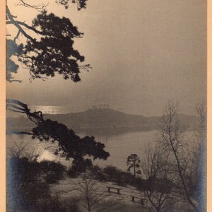 May include: A black and white photograph of a lake with a forested island in the middle. The lake is surrounded by trees and hills. The photo is taken from a high vantage point, looking down on the lake.