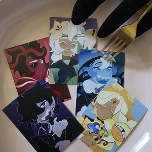 May include: Five rectangular stickers featuring stylized anime-style character illustrations in various colors including red, white, blue, and yellow. The stickers are arranged in a cluster, with a gold-toned metal fork resting on top of them.