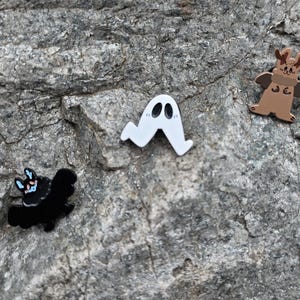 Puede incluir: Tres pines de esmalte con personajes de dibujos animados se muestran sobre una superficie de roca gris. Los pines incluyen un fantasma blanco, una criatura marrón con cuernos y alas, y un murciélago negro con detalles azules y rosas.