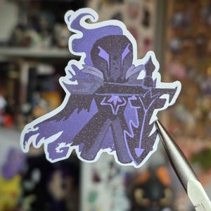 Op de afbeelding: Een sticker met een gestileerde ridder in paarse en zwarte tinten met glitteraccenten. De ridder draagt een helm en cape en houdt een zwaard en schild vast. De sticker heeft een witte rand en wordt vastgehouden door een gereedschap.