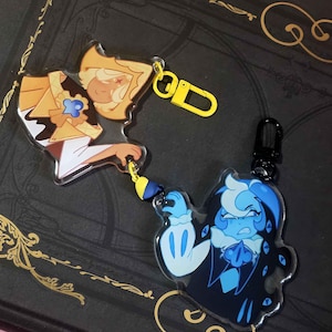 Pode incluir: Dois chaveiros de acrílico com personagens de desenhos animados. Um chaveiro tem um personagem amarelo e marrom com um fecho amarelo. O outro chaveiro tem um personagem azul e preto com um fecho preto. Ambos em um fundo escuro.