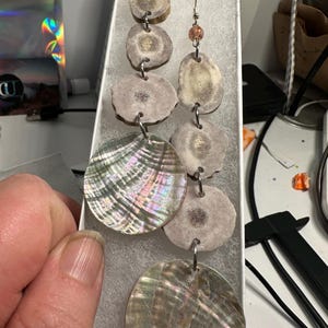 May include: Handmade earrings featuring a combination of natural materials. The earrings have a series of round, light-colored antler pieces and iridescent, round shell pendants. The earrings are connected with silver-colored metal links and have orange-colored bead accents.