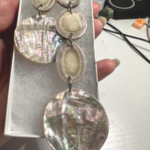 May include: Handmade earrings featuring a combination of natural antler slices and iridescent shell pendants. The earrings have a unique, organic design with a mix of neutral and shimmering colors, suspended by silver-tone metal rings.