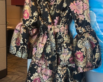 May include: A black coat with a floral pattern in gold, pink, and green. The coat has large bell sleeves and a cinched waist. The fabric has a brocade texture. The coat is hanging on a blue hanger.