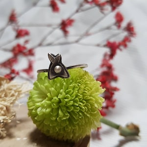 May include: A silver ring with a small, triangular design featuring a pearl in the center. The ring is resting on a green flower.