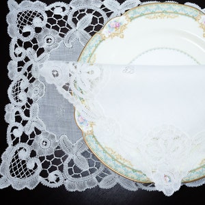 May include: A white lace doily with a white porcelain plate and a white linen napkin folded inside. The plate has a gold rim and a floral design.