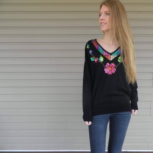 Könnte beinhalten: Ein schwarzer Pullover mit einem bunten Paillettenmuster am Ausschnitt. Das Muster zeigt eine rosa Blume, grüne Blätter und schimmernde Pailletten.