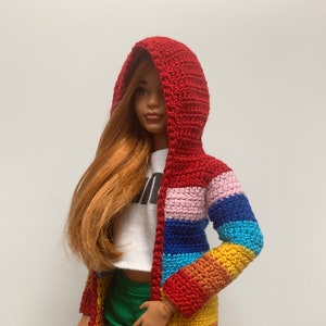 Puede incluir: Una muñeca con un suéter con capucha rojo tejido a ganchillo con un diseño de rayas arcoíris. El suéter está abierto por delante y tiene mangas largas. La muñeca también lleva una camiseta blanca y unos pantalones cortos verdes.