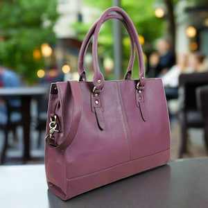 May include: A structured plum-coloured leather handbag featuring two top handles, a detachable shoulder strap with a metal lobster clasp, and vertical stitched detailing on the front panel. The bag has a rectangular base and antique brass hardware.