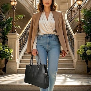 Peut inclure: Un blazer beige porté sur un chemisier blanc croisé et un jean bleu clair. La tenue est complétée par un sac fourre-tout noir structuré doté de deux anses et de détails argentés, tenu sur le côté dans un ensemble professionnel.