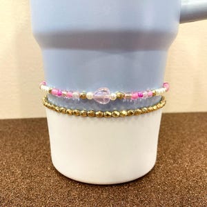 May include: A light blue ceramic mug with a white band around the middle. The band is decorated with two rows of beads, one row of gold beads and one row of pink, white, and gold beads with a purple heart-shaped bead.