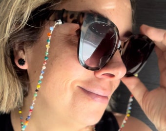 Puede incluir: Gafas de sol con montura de carey y una cadena de cuentas multicolores. La cadena presenta pequeñas cuentas redondas de varios colores. Las gafas de sol tienen lentes oscuras y una forma de montura clásica.