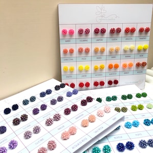 Może przedstawiać: Wystawa małych, okrągłych, kolorowych kolczyków z koralików. Kolczyki są ułożone w rzędach na białej kartce z nadrukowanymi pod nimi włoskimi nazwami każdego koloru. Kartka nosi tytuł "Gisele da indossare", co tłumaczy się jako "Prezent do noszenia".