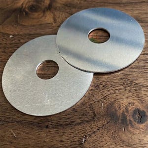 May include: Two circular, flat metal washers with a brushed silver finish. Each piece features a centered, circular hole. The surfaces are smooth with a metallic sheen, showing a consistent, plain design intended for hardware or craft applications.