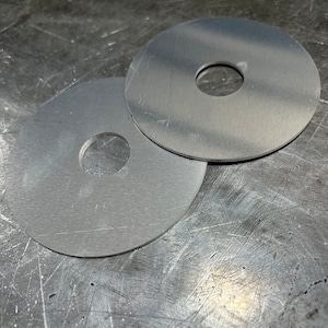 May include: Two silver metal washers with a hole in the center. The washers are on a scratched metal surface.