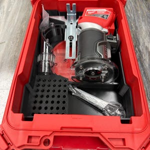 May include: A red toolbox with a clear lid, containing a black and red power router, accessories, and a black storage tray. The router has a black handle and a clear base. The toolbox is open, revealing the tools inside.