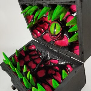 Puede incluir: Un cofre del tesoro de madera negra, abierto para revelar un interior monstruoso. El interior presenta superficies rojas texturizadas, dientes verdes y puntiagudos, y un gran ojo verde con una pupila en forma de hendidura amarilla. El cofre se cierra con un cierre de metal.
