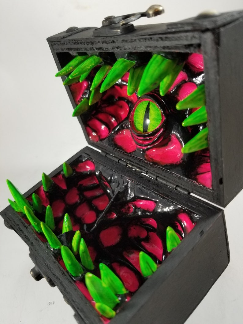 Puede incluir: Un cofre del tesoro de madera negra, abierto para revelar un interior monstruoso. El interior presenta superficies rojas texturizadas, dientes verdes y puntiagudos, y un gran ojo verde con una pupila en forma de hendidura amarilla. El cofre se cierra con un cierre de metal.