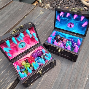 Puede incluir: Dos cofres del tesoro de madera abiertos con un tema de fantas&iacute;a. Los interiores est&aacute;n pintados con un dise&ntilde;o de boca de monstruo, con dientes y un ojo. Los cofres est&aacute;n llenos de dados de colores.