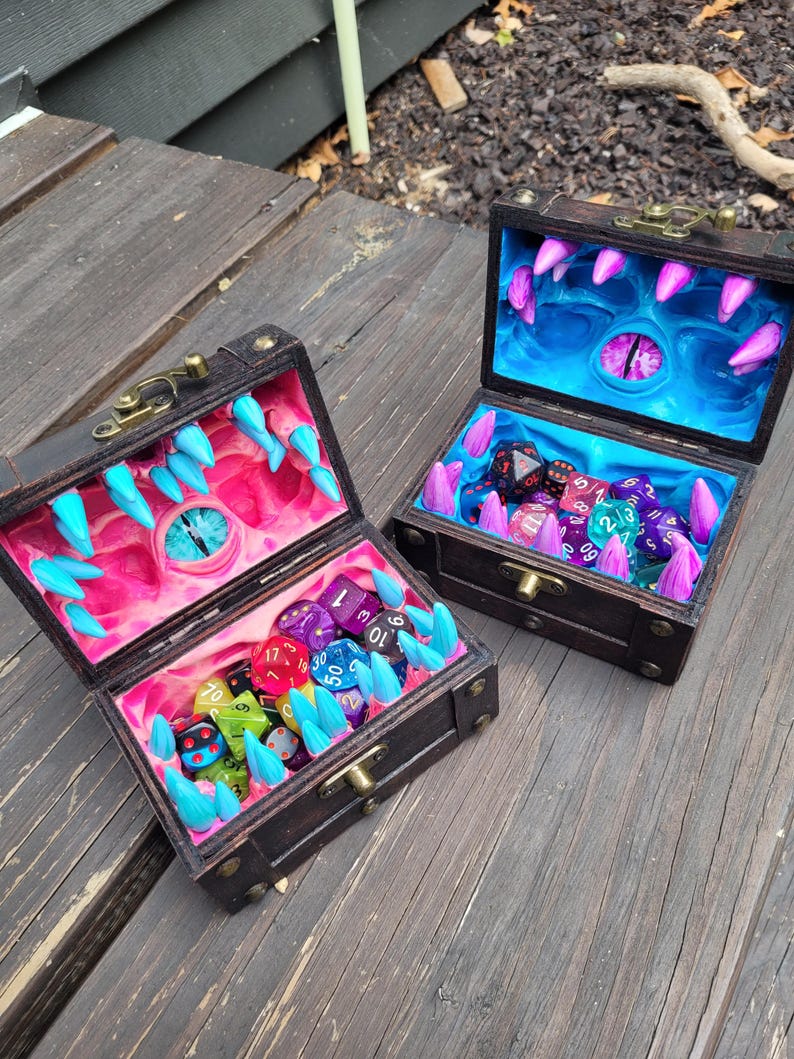 Puede incluir: Dos cofres del tesoro de madera abiertos con un tema de fantas&iacute;a. Los interiores est&aacute;n pintados con un dise&ntilde;o de boca de monstruo, con dientes y un ojo. Los cofres est&aacute;n llenos de dados de colores.